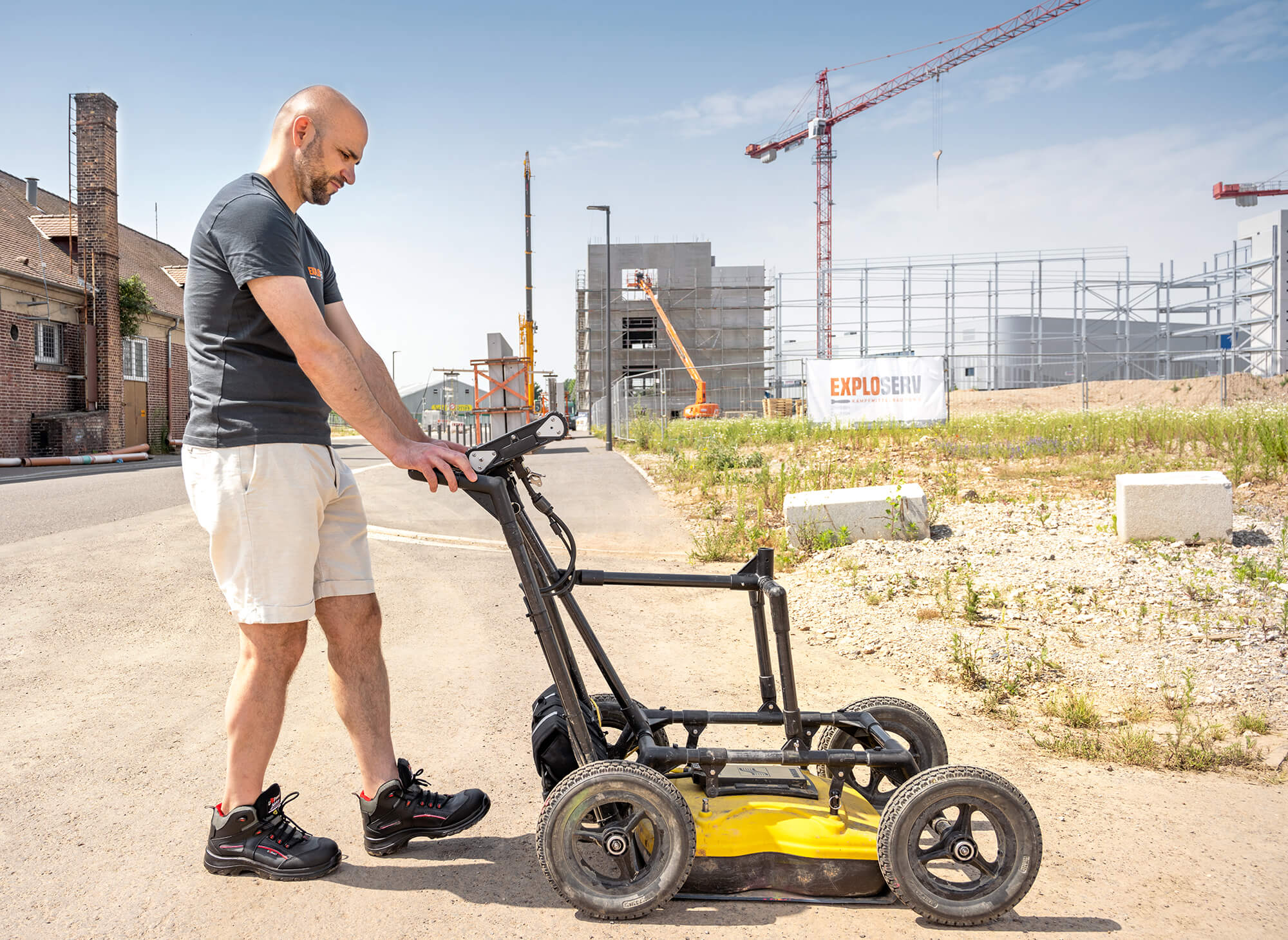 Ein Mann in kurzer Arbeitshose und T-Shirt schiebt ein Bodenradar-Gerät über einen befestigten Weg. Im Hintergrund sind Baustellen, Kräne und moderne Gebäude unter blauem Himmel zu sehen.“