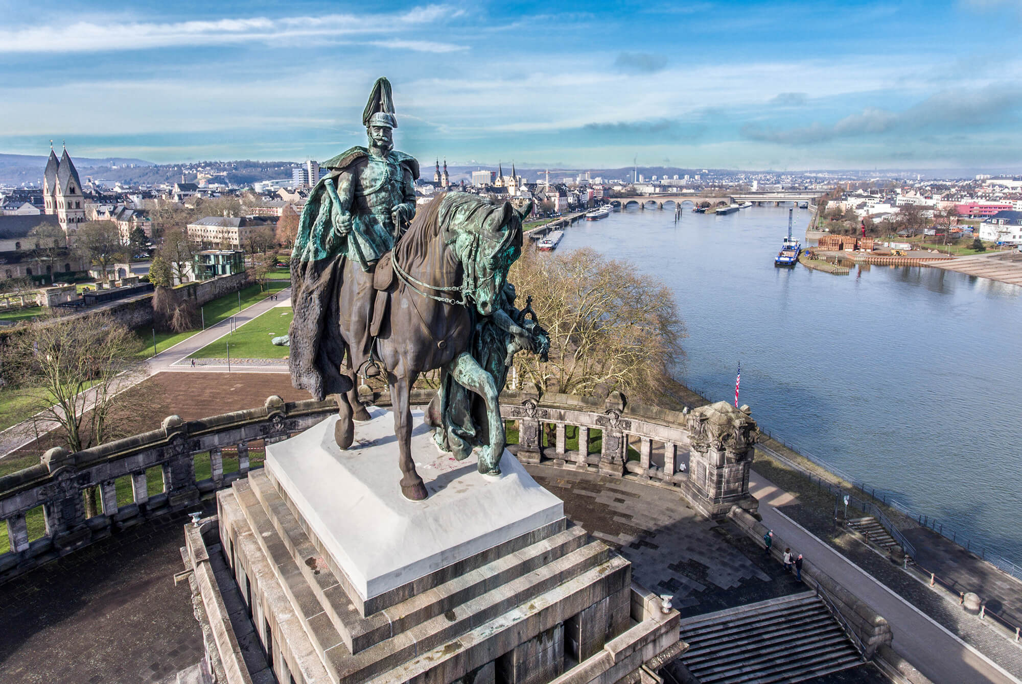 Luftaufnahme eines monumentalen Reiterdenkmals am Zusammenfluss von Rhein und Mosel. Im Hintergrund sind die Stadt, Uferpromenaden und Brücken unter blauem Himmel zu sehen.
