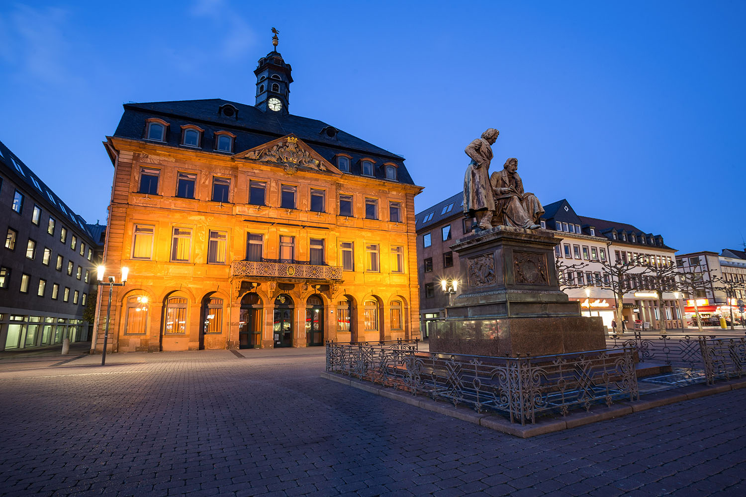 Abendaufnahme eines historischen Gebäudes mit Turmuhr, warm beleuchtet im Zentrum einer Stadt. Im Vordergrund steht eine Statue auf einem Brunnen, umgeben von Kopfsteinpflaster und Straßenlaternen.