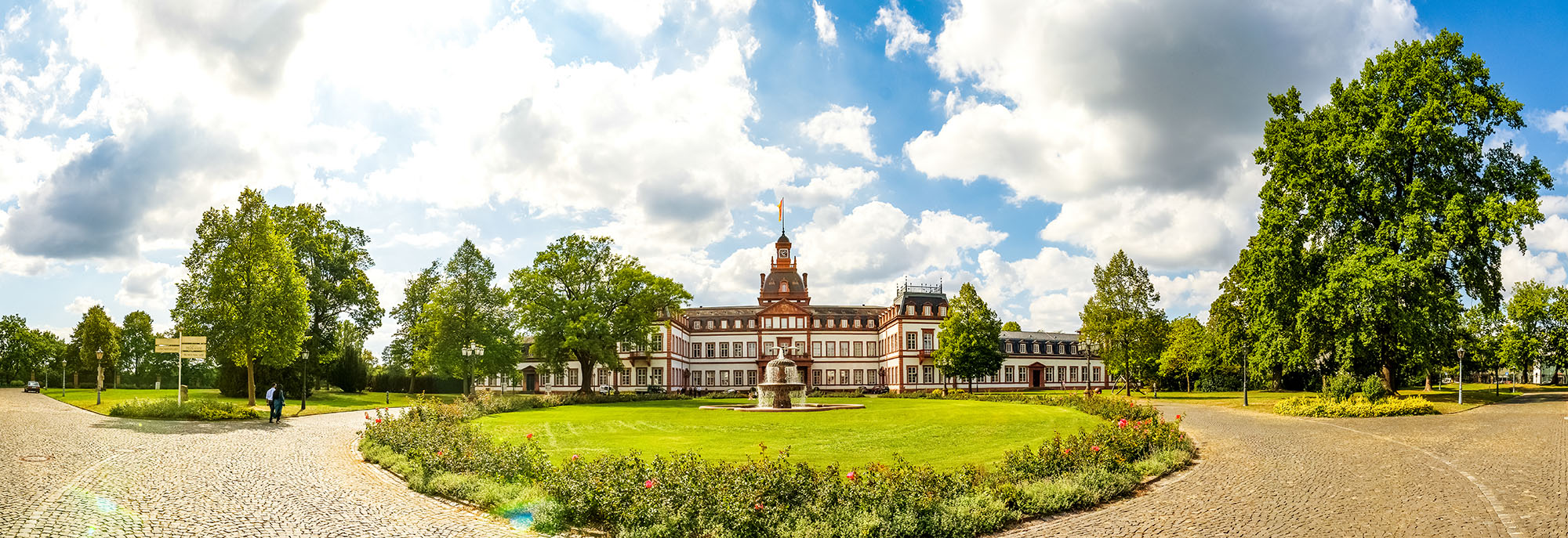 Panoramaaufnahme einer historischen Parkanlage mit symmetrisch angelegten Wegen und einem zentralen Schlossgebäude im Hintergrund. Umgeben von gepflegten Grünflächen, Bäumen und blauem Himmel mit weißen Wolken.