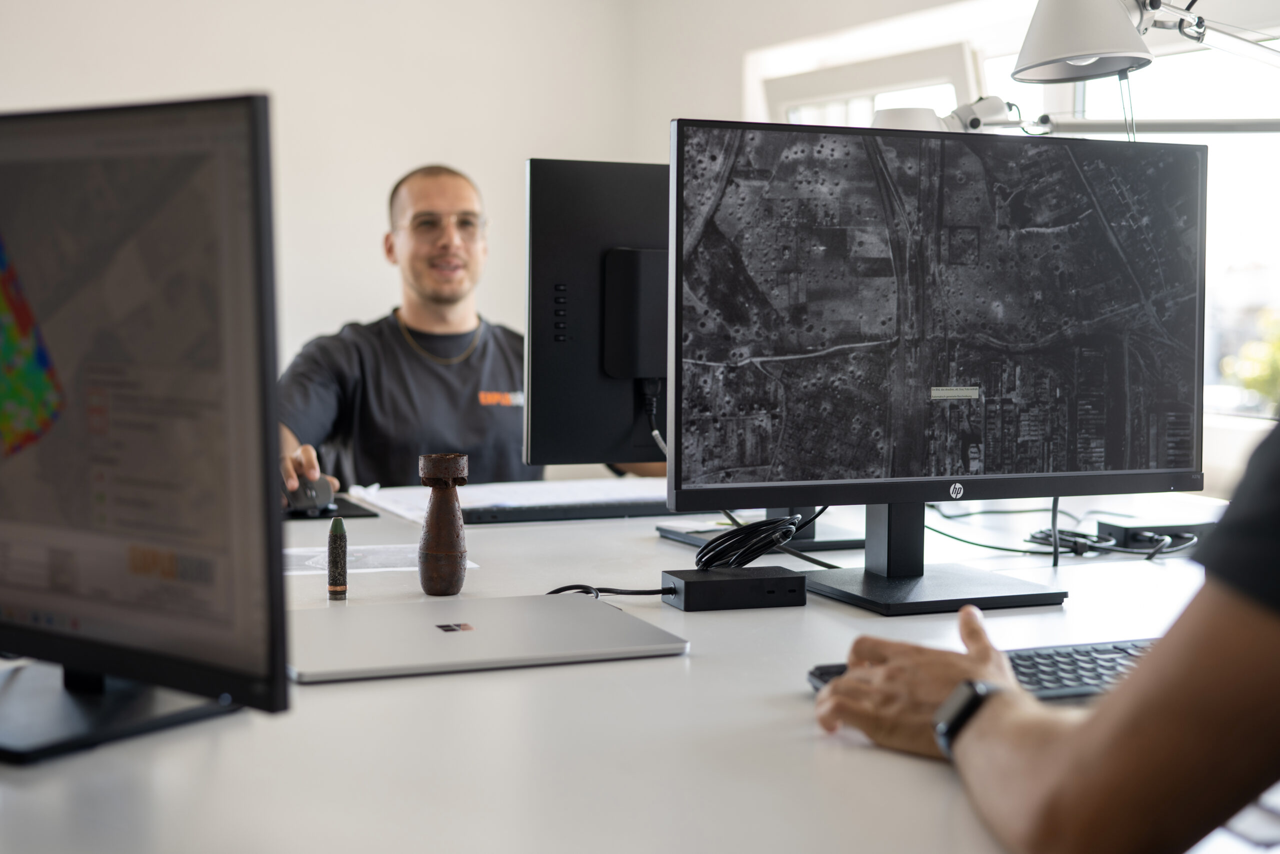 Zwei Mitarbeiter sitzen an Schreibtischen und analysieren digitale Karten auf großen Monitoren. Im Vordergrund ist ein Satellitenbild zu sehen, im Hintergrund ein Mitarbeiter mit grauem ‚Exploserv‘-Shirt.