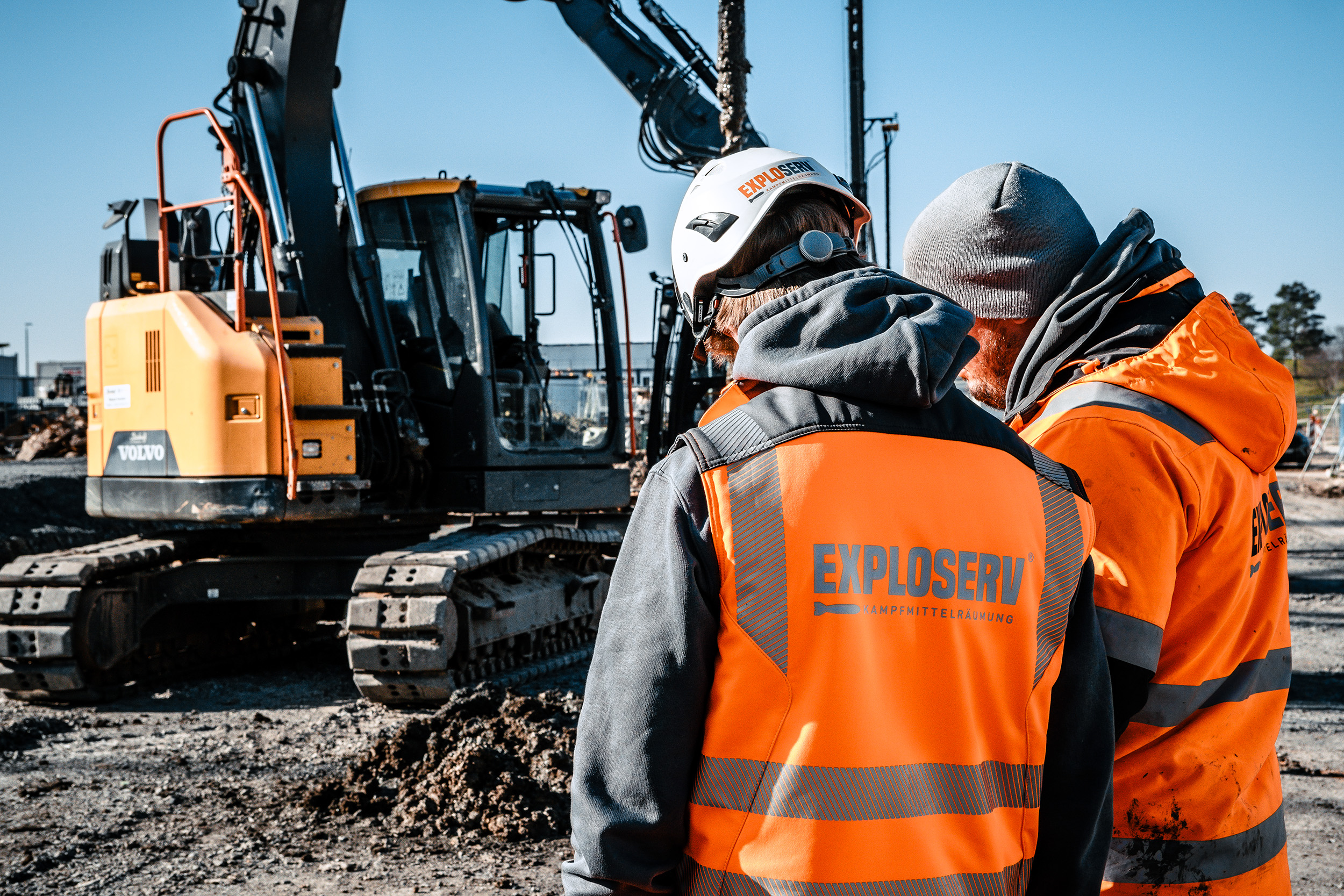 Zwei Arbeiter mit Schutzhelmen und orangefarbenen Warnwesten mit dem Logo ‚Exploserv‘ besprechen sich auf einer Baustelle. Im Hintergrund steht ein gelber Bagger auf geschottertem Boden.