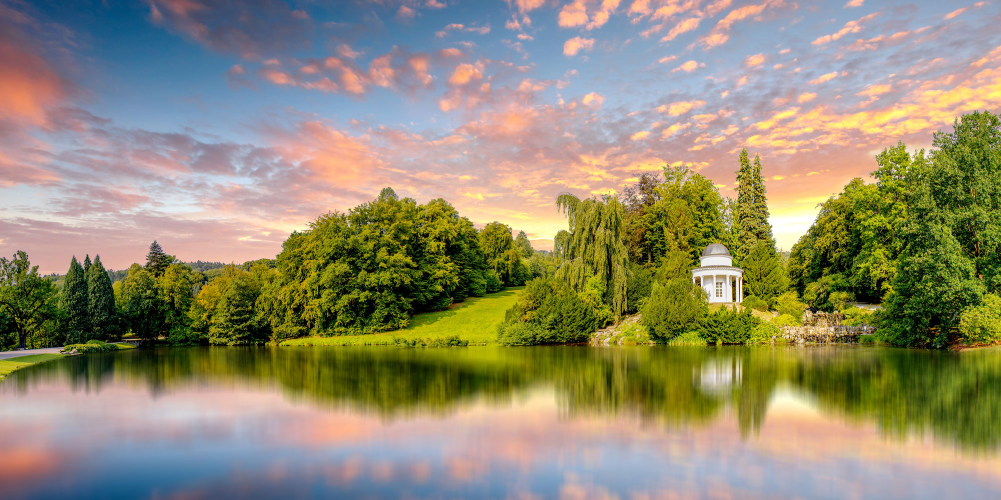 Idyllische Landschaft mit einem kleinen weißen Pavillon am Ufer eines Sees, umgeben von dichtem Grün. Der Himmel leuchtet in warmen Farben des Sonnenuntergangs und spiegelt sich im ruhigen Wasser wider.