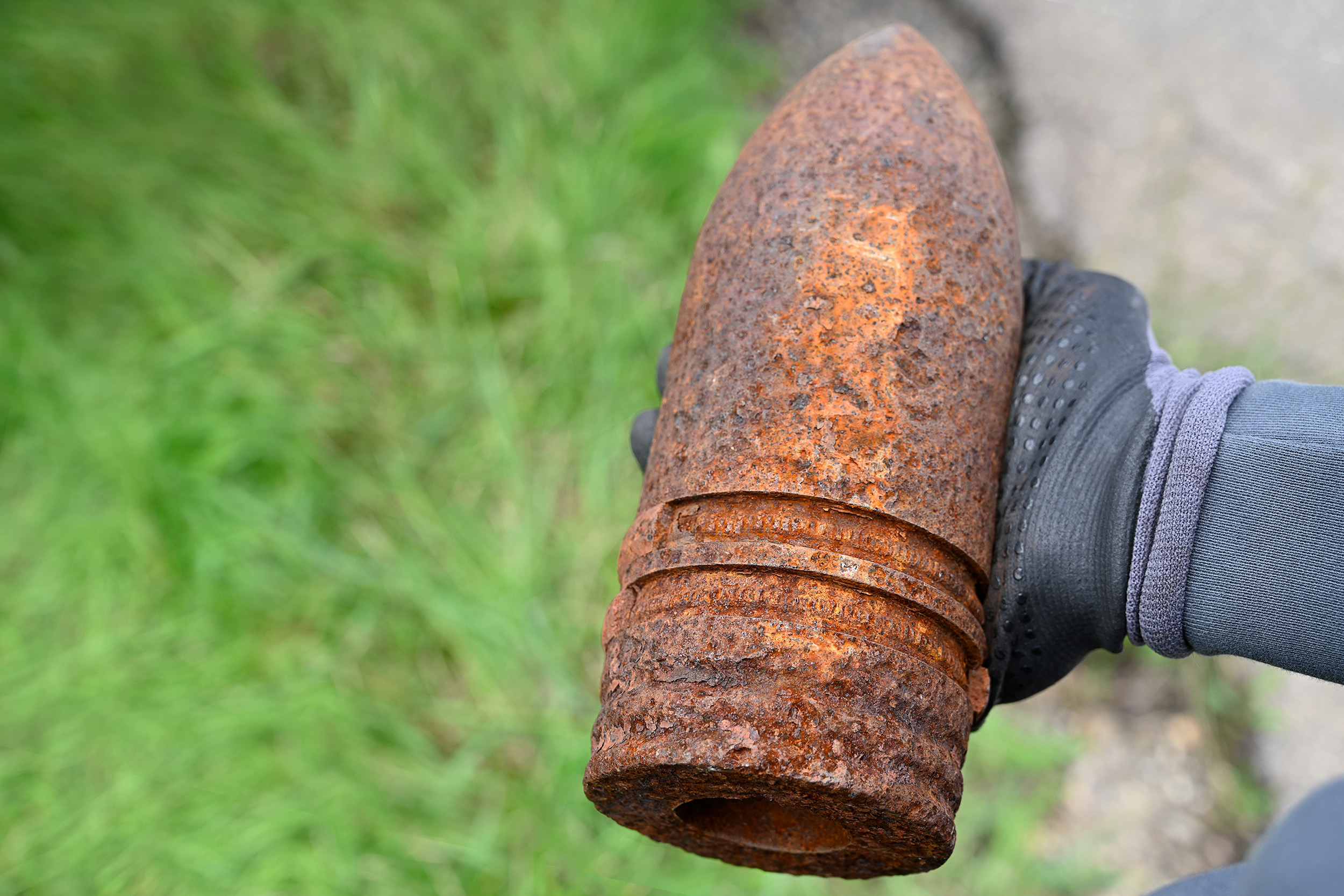 Eine Hand mit Schutzhandschuh hält eine stark verrostete alte Munition oder Granate vor grünem Hintergrund.