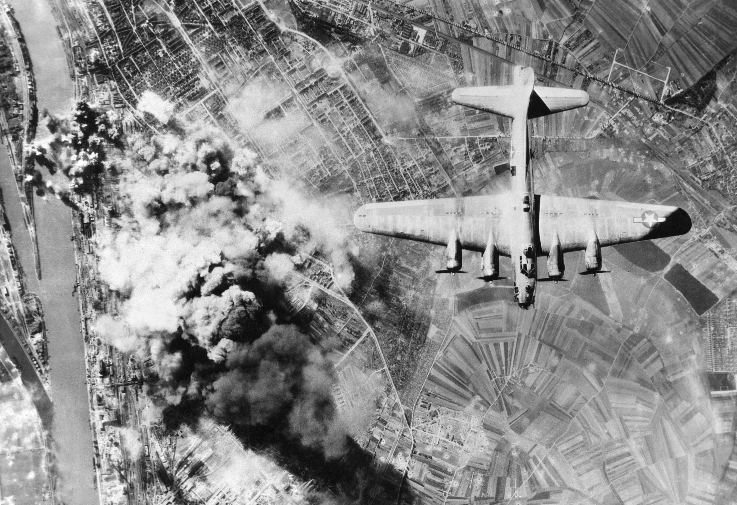 Schwarz-Weiß-Luftaufnahme eines Bombenabwurfs während eines historischen Luftangriffs. Ein großes Flugzeug fliegt über eine Landschaft, während unter ihm Explosionen und Rauchwolken aufsteigen.