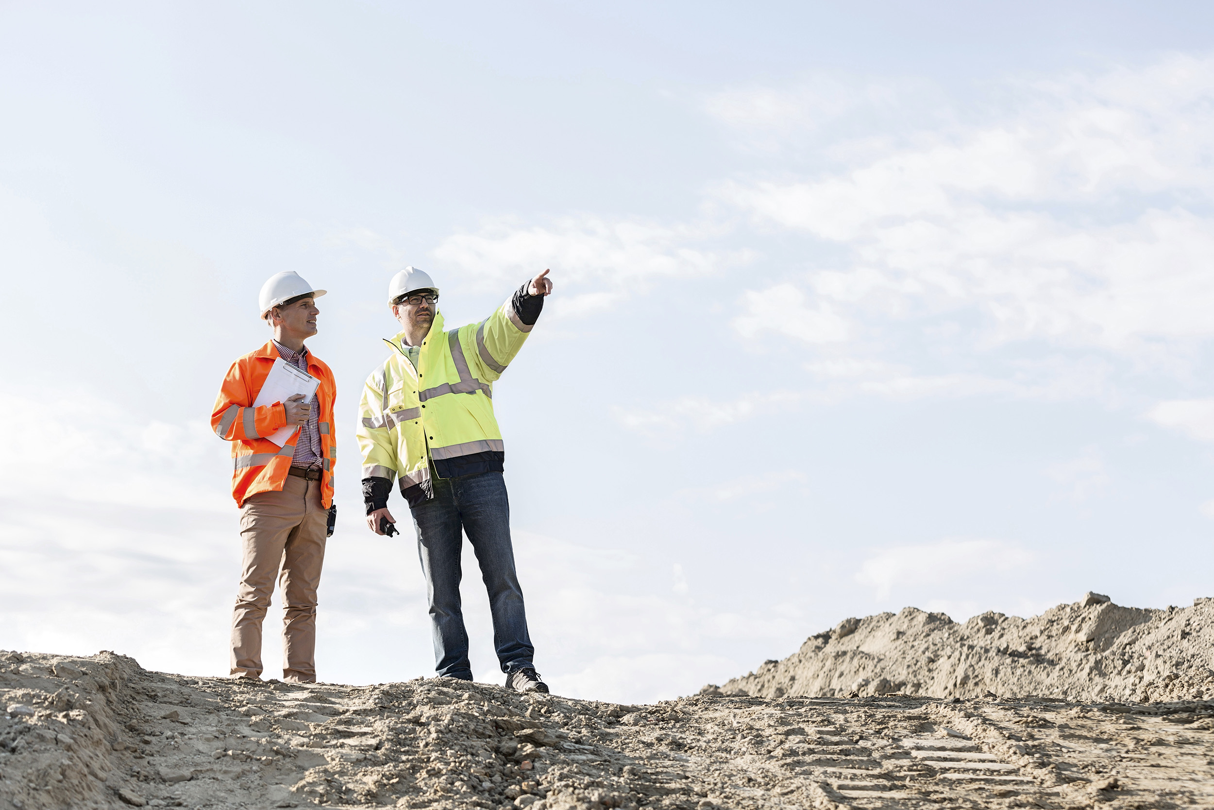 Zwei Personen mit Schutzhelmen und Warnwesten stehen auf einer Baustelle und blicken in die Ferne, während eine von ihnen in eine bestimmte Richtung zeigt.