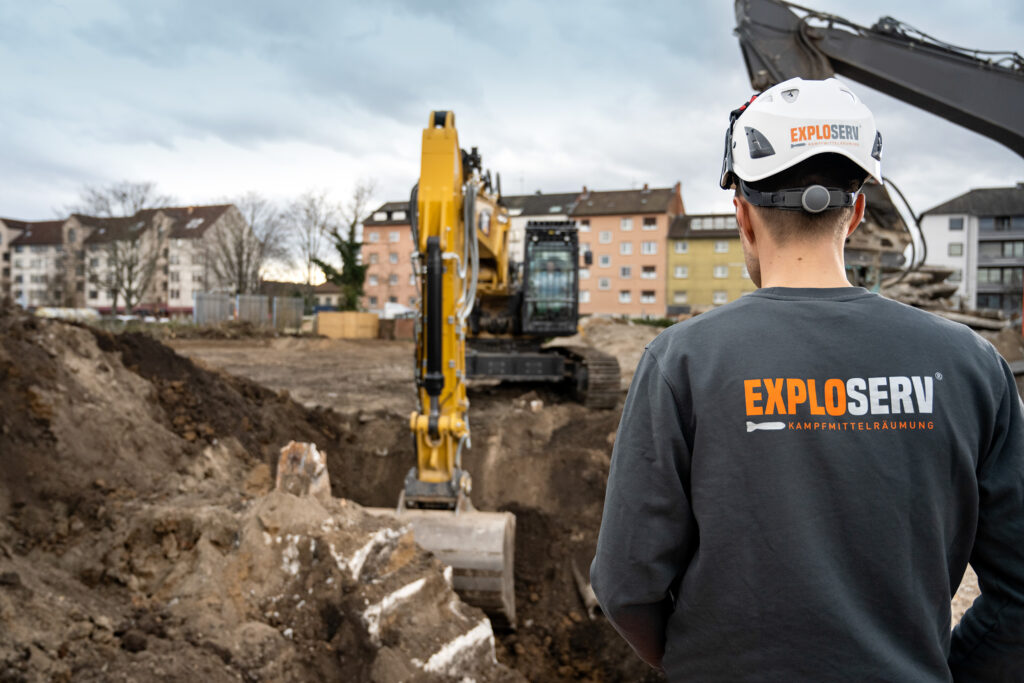 Ein Mitarbeiter mit Schutzhelm und grauem ‚Exploserv‘-Shirt beobachtet einen Bagger, der auf einer Baustelle Erdreich aushebt. Im Hintergrund stehen Wohnhäuser unter bewölktem Himmel.
