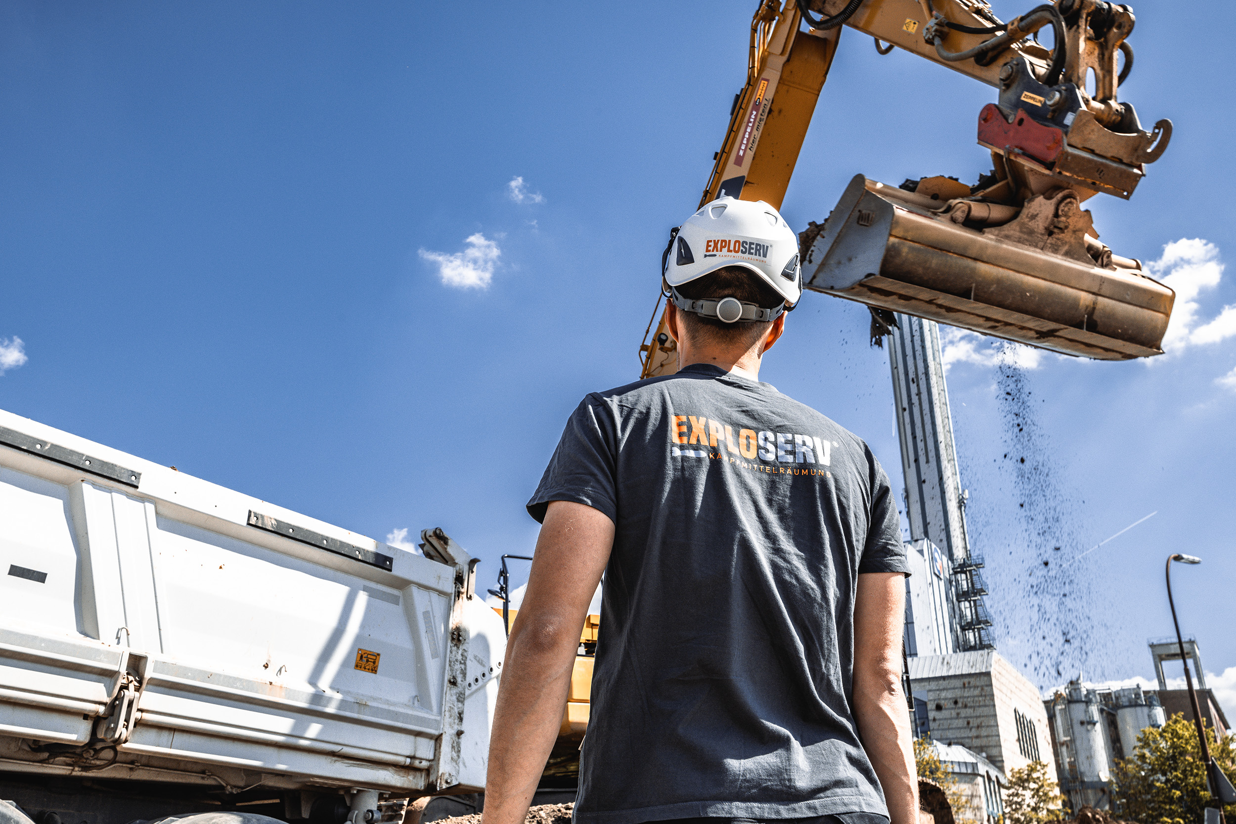 Ein Bauarbeiter mit Schutzhelm und grauem Firmen-T-Shirt steht auf einer Baustelle und beobachtet einen Bagger, der gerade einen schweren Holzstamm hebt. Im Hintergrund sind ein Lkw, Baukräne und ein klarer blauer Himmel zu sehen.“