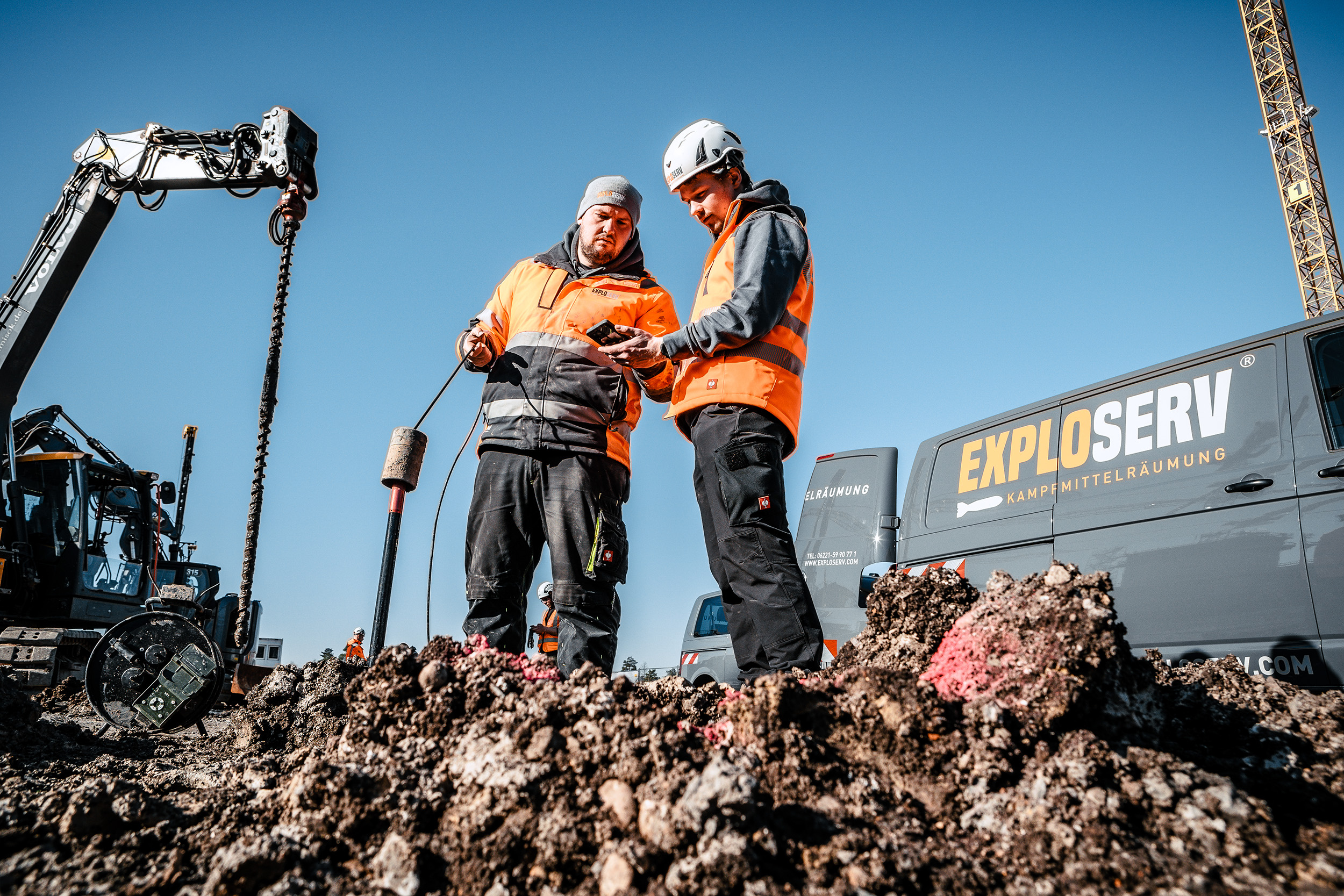 Zwei Arbeiter mit orangefarbenen Warnwesten und Schutzhelmen stehen auf einer Baustelle und besprechen Messdaten. Im Hintergrund sind ein Bagger, Maschinen und ein Container mit der Aufschrift ‚Exploserv‘ zu sehen, unter einem klaren blauen Himmel.