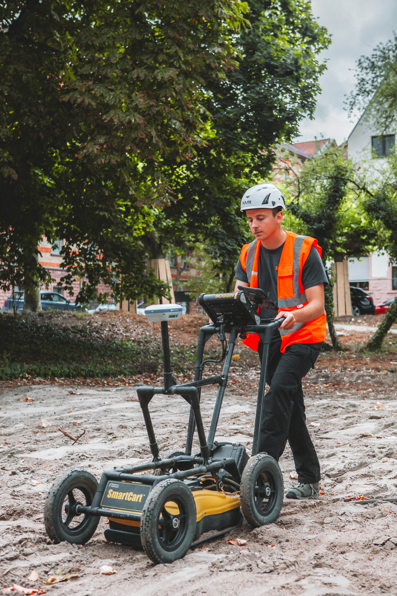 Ein Mitarbeiter mit Schutzhelm und orangefarbener Warnweste bedient ein Bodenradar-Gerät auf einem freigeräumten Gelände. Hinter ihm stehen Bäume und Gebäude in einer städtischen Umgebung.