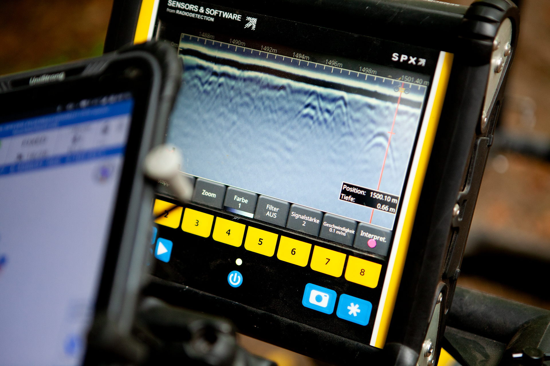 Nahaufnahme eines digitalen Messgeräts mit gelbem Rahmen, das ein Scanbild auf dem Display zeigt. Daneben ist teilweise ein zweites Gerät mit blauer Benutzeroberfläche zu sehen.