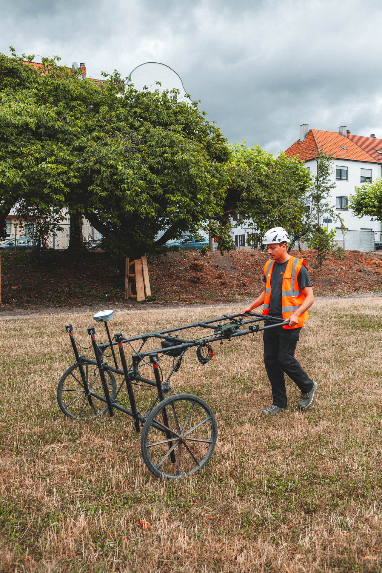 Ein Mitarbeiter mit orangefarbener Warnweste und Schutzhelm führt eine manuelle Messung mit einem Metallsuchgerät auf einem freigeräumten Gelände durch. Im Hintergrund sind Bäume und geparkte Fahrzeuge zu sehen.“