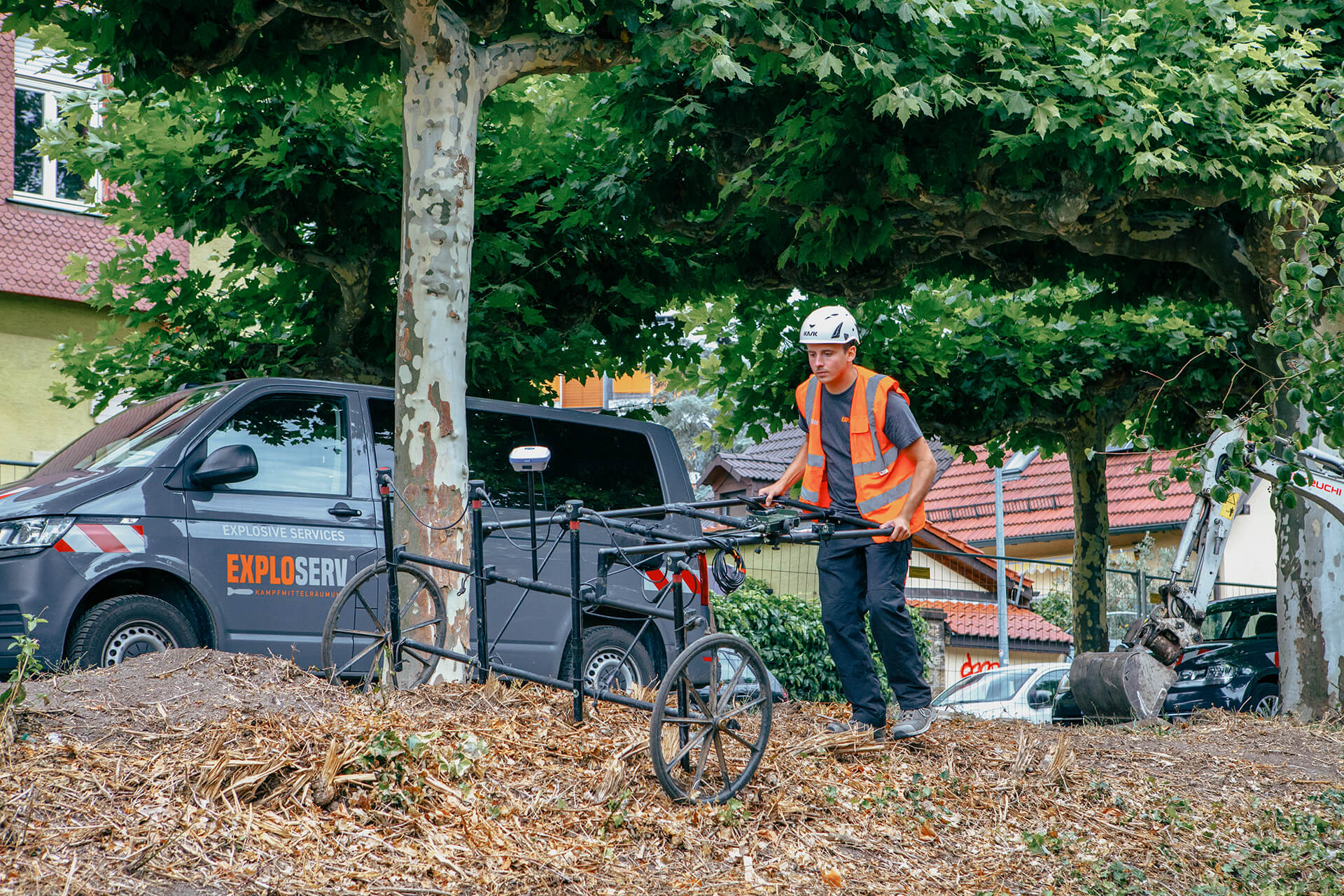 Ein Mitarbeiter mit Schutzhelm und orangefarbener Warnweste führt ein Bodenradar-Gerät durch ein mit Erde bedecktes Gebiet. Im Hintergrund steht ein grauer Firmenwagen mit dem Logo ‚Exploserv‘ unter Bäumen.