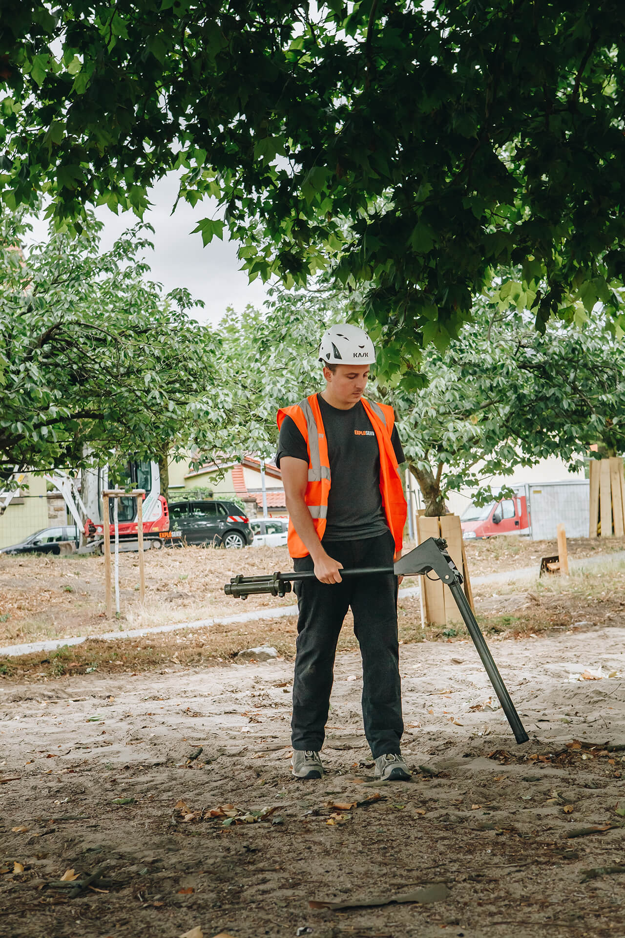 Ein Mitarbeiter mit orangefarbener Warnweste und Schutzhelm führt eine manuelle Messung mit einem Metallsuchgerät auf einem freigeräumten Gelände durch. Im Hintergrund sind Bäume und geparkte Fahrzeuge zu sehen.