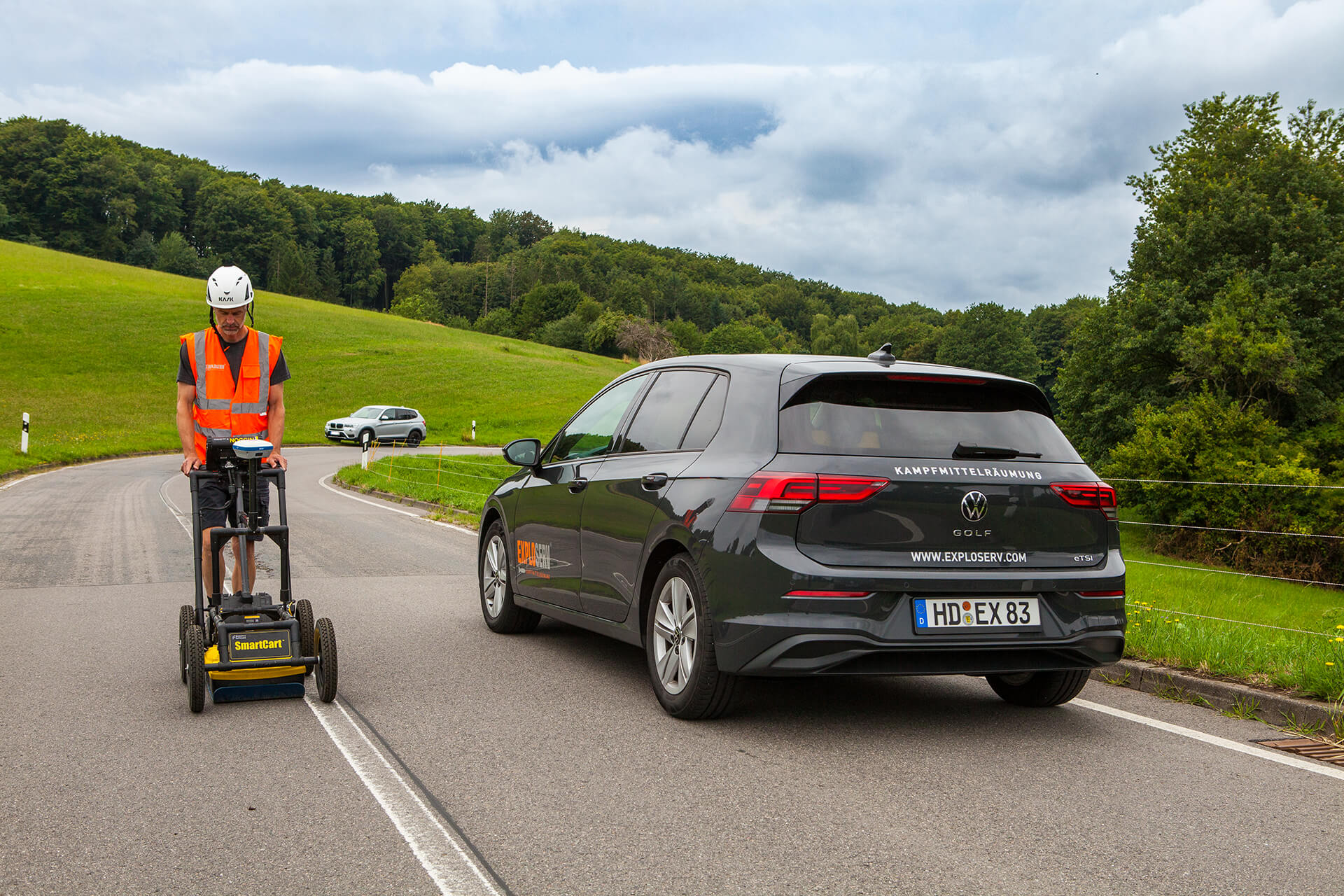 Ein Mitarbeiter mit orangefarbener Warnweste und Schutzhelm führt eine manuelle Messung mit einem Metallsuchgerät auf einem freigeräumten Gelände durch. Im Hintergrund sind Bäume und geparkte Fahrzeuge zu sehen.“