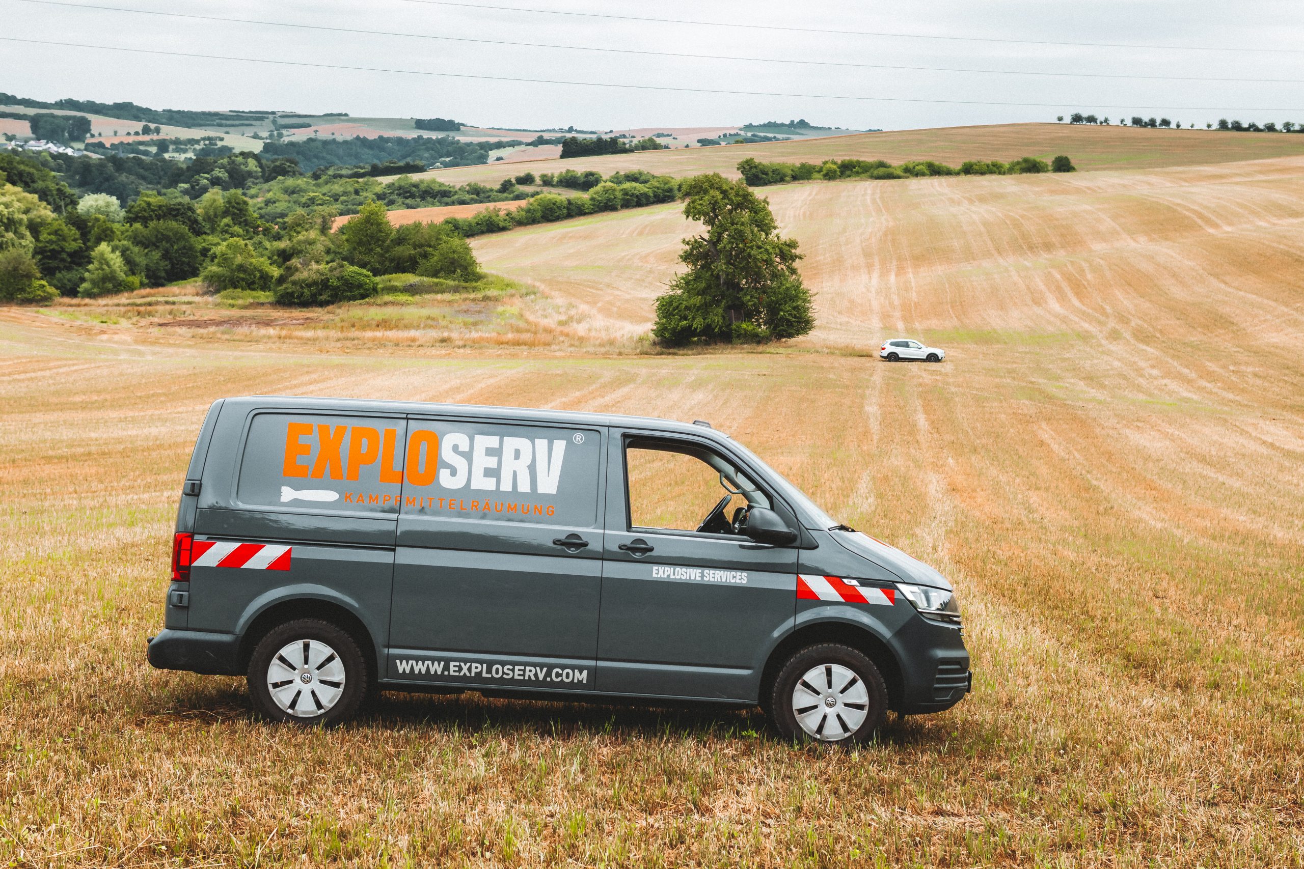 Ein grauer Firmenwagen mit dem Logo ‚Exploserv‘ steht auf einem abgeernteten Feld in hügeliger Landschaft. Im Hintergrund sind Bäume, Wiesen und ein weiteres Fahrzeug in der Ferne zu sehen.