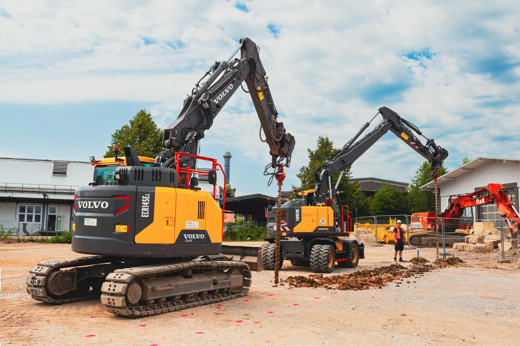 Zwei gelbe Bagger arbeiten nebeneinander auf einer Baustelle. Einer hebt Erde aus, während der andere Material ablädt. Im Hintergrund stehen Industriehallen und Bäume unter leicht bewölktem Himmel.