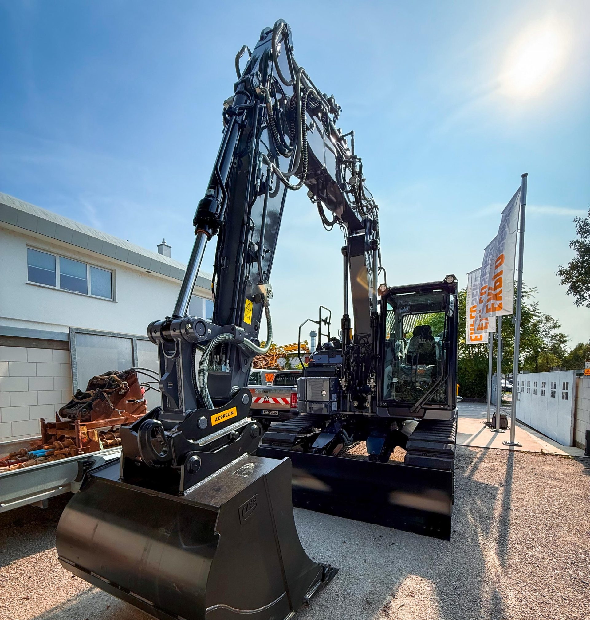 Ein schwarzer Kettenbagger steht im Sonnenlicht auf einem Firmengelände. Der Baggerarm ist leicht angehoben, im Hintergrund sind Gebäude und eine Firmenfahne zu sehen.