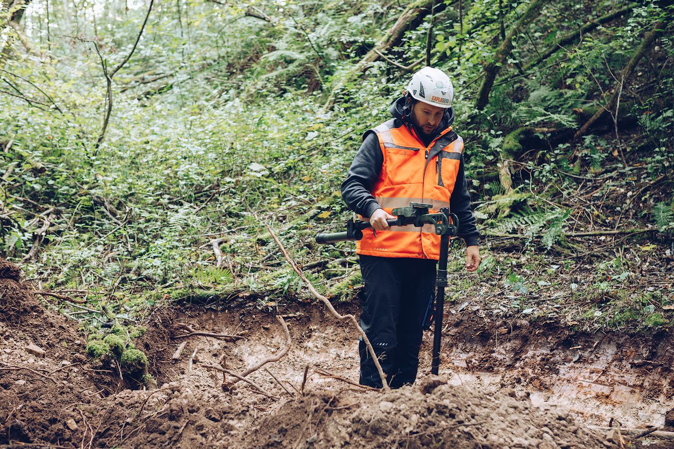 Eine Person mit orangefarbener Warnweste und Schutzhelm untersucht mit einem Messgerät den Boden einer Grabungsstelle im Wald.
