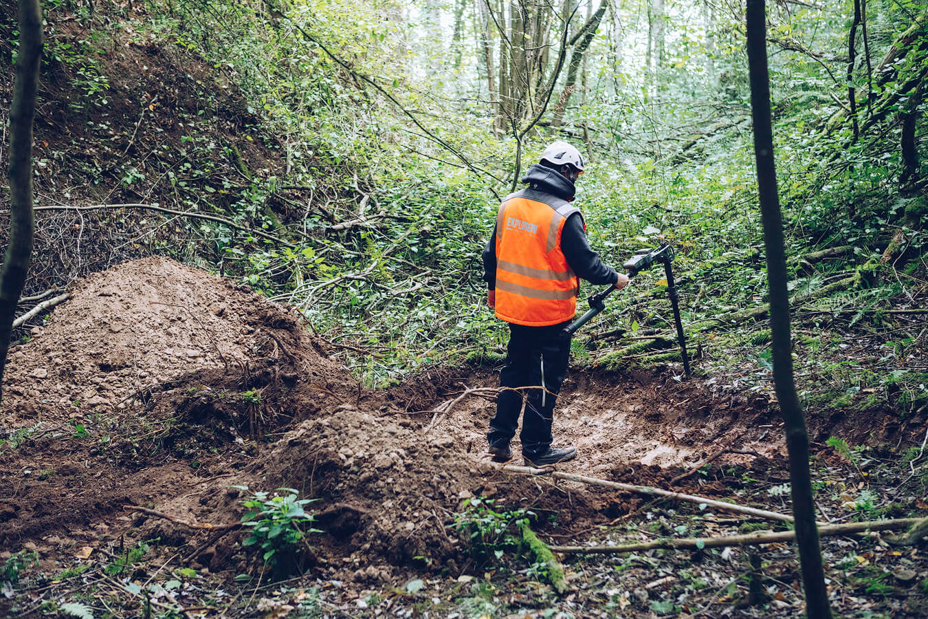 Eine Person mit Schutzhelm und orangefarbener Warnweste steht in einer bewaldeten Grabungsstelle und untersucht den Boden mit einem Messgerät.