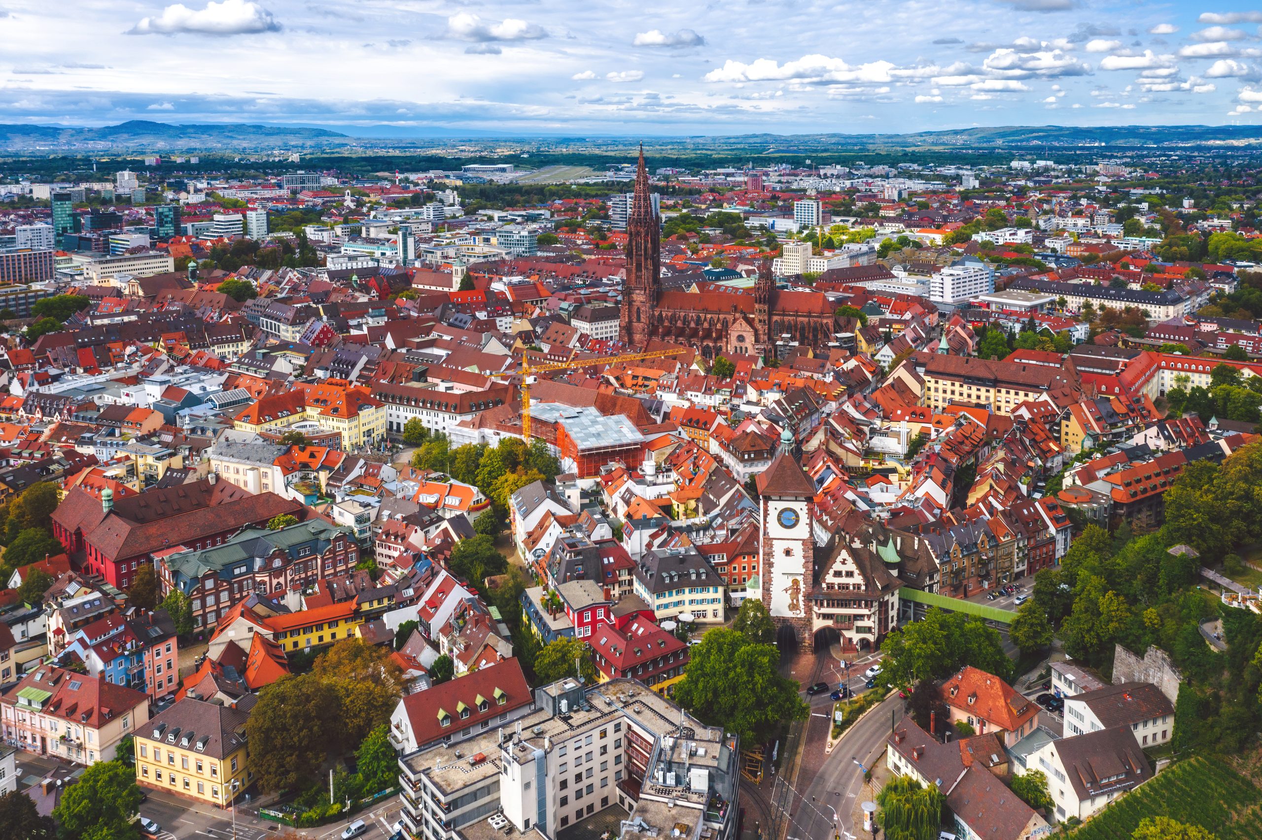 Luftaufnahme der Innenstadt von Freiburg im Breisgau mit dem Freiburger Münster im Zentrum, umgeben von roten Dächern und historischen Gebäuden. Im Hintergrund ist die Stadt mit dem Schwarzwald zu sehen.“