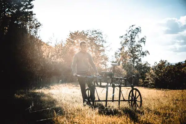 „Person steht auf einer Wiese und führt ein technisches Messgerät auf Rädern, umgeben von Sonnenlicht und Bäumen im Hintergrund.