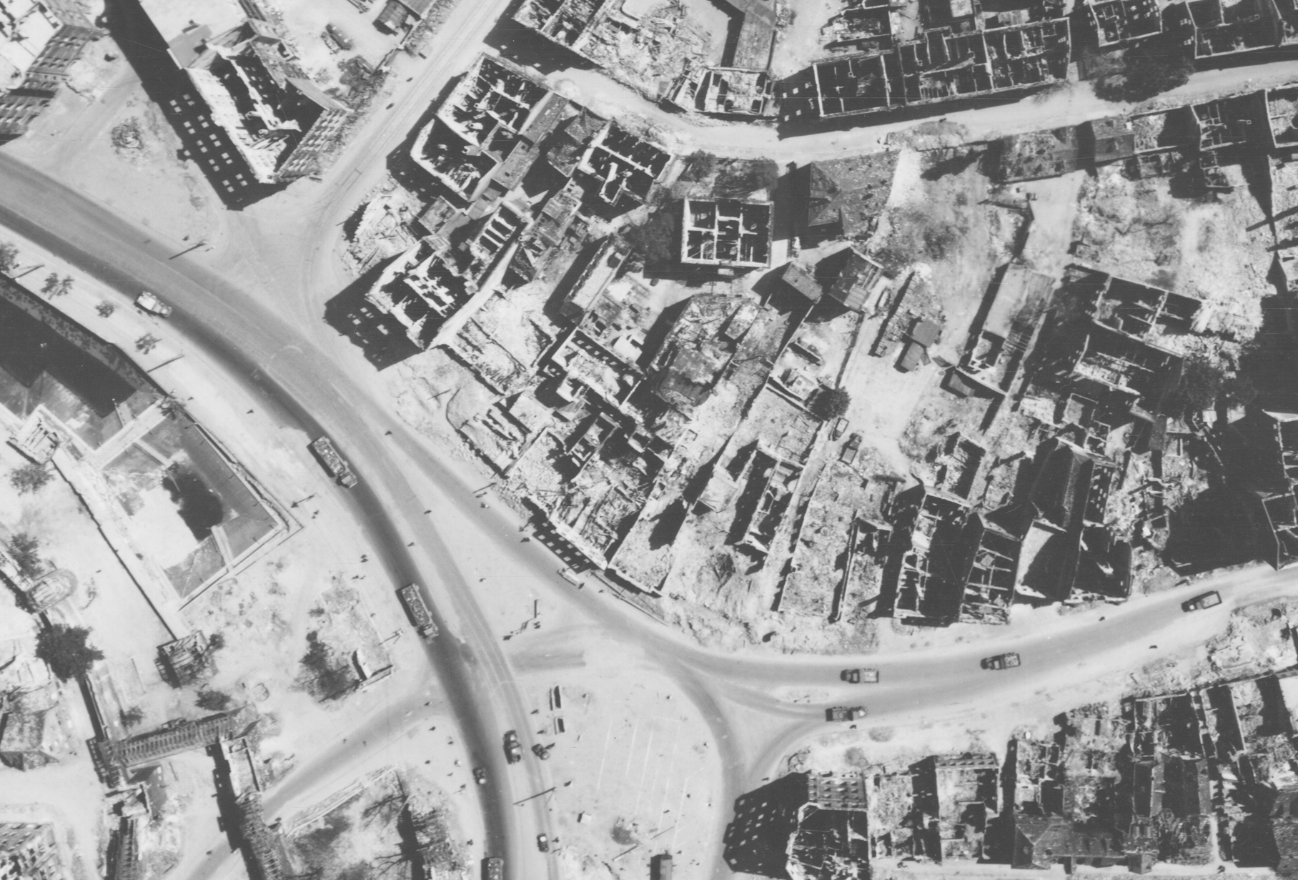 Schwarz-Weiß-Luftaufnahme einer zerstörten Stadt nach einem Luftangriff. Zu erkennen sind Straßenzüge, Trümmer und beschädigte Gebäude, die das Ausmaß der Zerstörung zeigen.