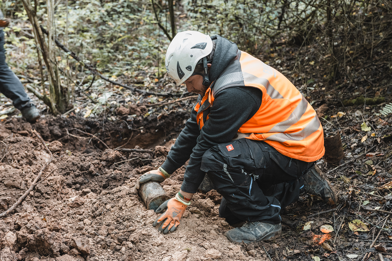 Eine Person mit Schutzhelm und orangefarbener Warnweste kniet auf einer Grabungsstelle im Wald und legt vorsichtig einen metallischen Gegenstand im Erdreich frei.