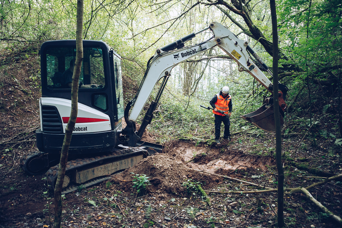Ein Bagger hebt Erde in einem bewaldeten Gebiet aus, während eine Person in Warnweste und Schutzhelm die Grabungsstelle kontrolliert.