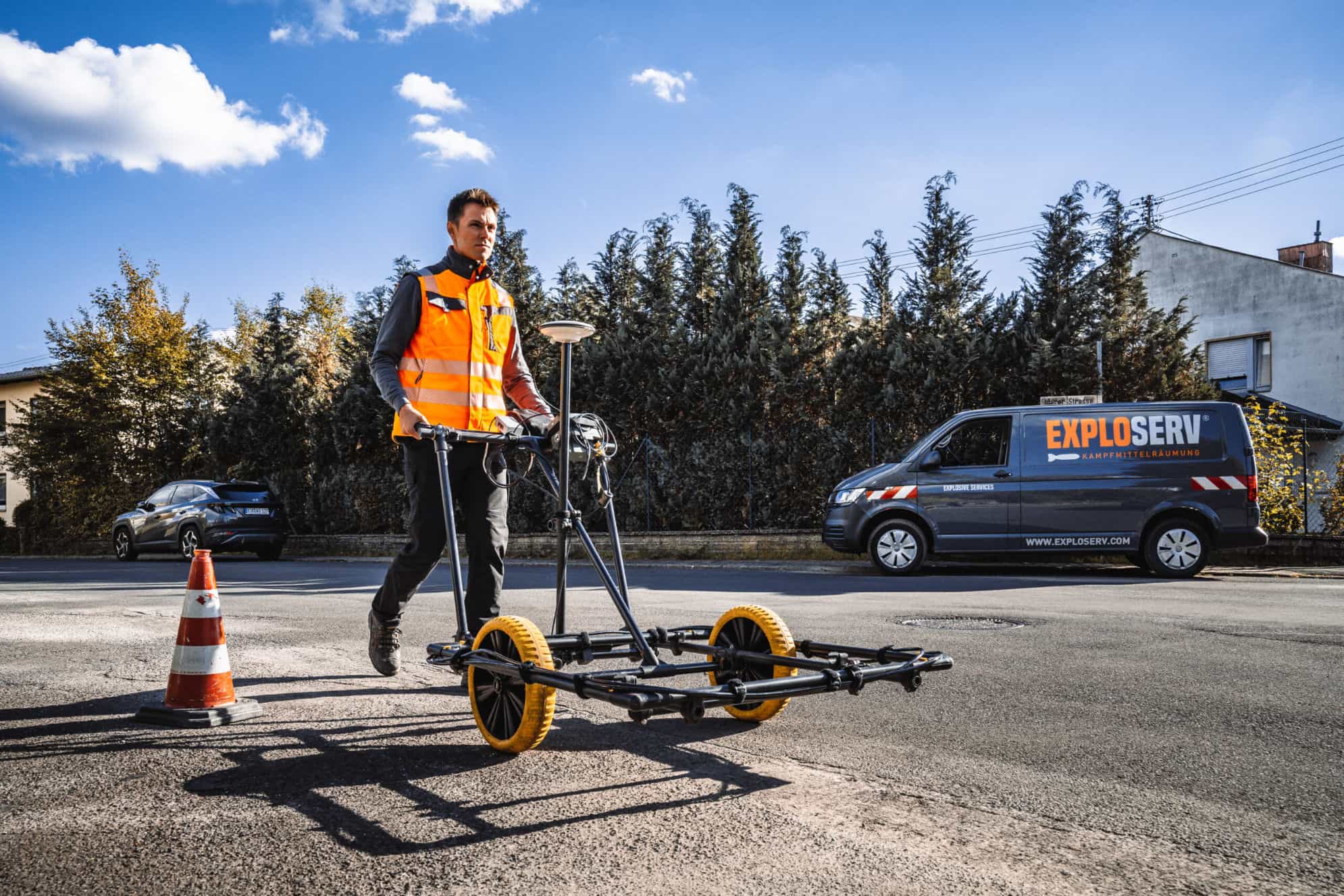 Mitarbeiter in orangefarbener Warnweste schiebt auf einer Straße ein Messgerät zur Kampfmittelsondierung, im Hintergrund ein Firmenfahrzeug von Exploserv.