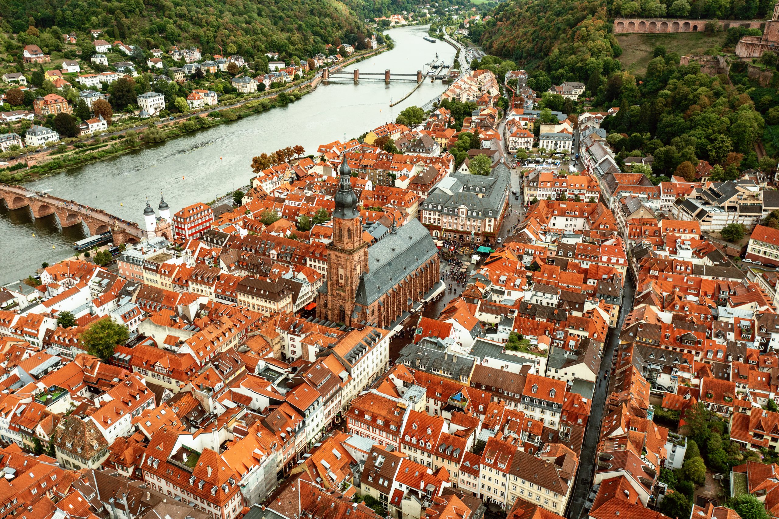 Luftaufnahme der Altstadt von Heidelberg mit ihren roten Dächern und der Heiliggeistkirche im Zentrum. Im Hintergrund fließt der Neckar mit der Alten Brücke, eingerahmt von bewaldeten Hügeln.