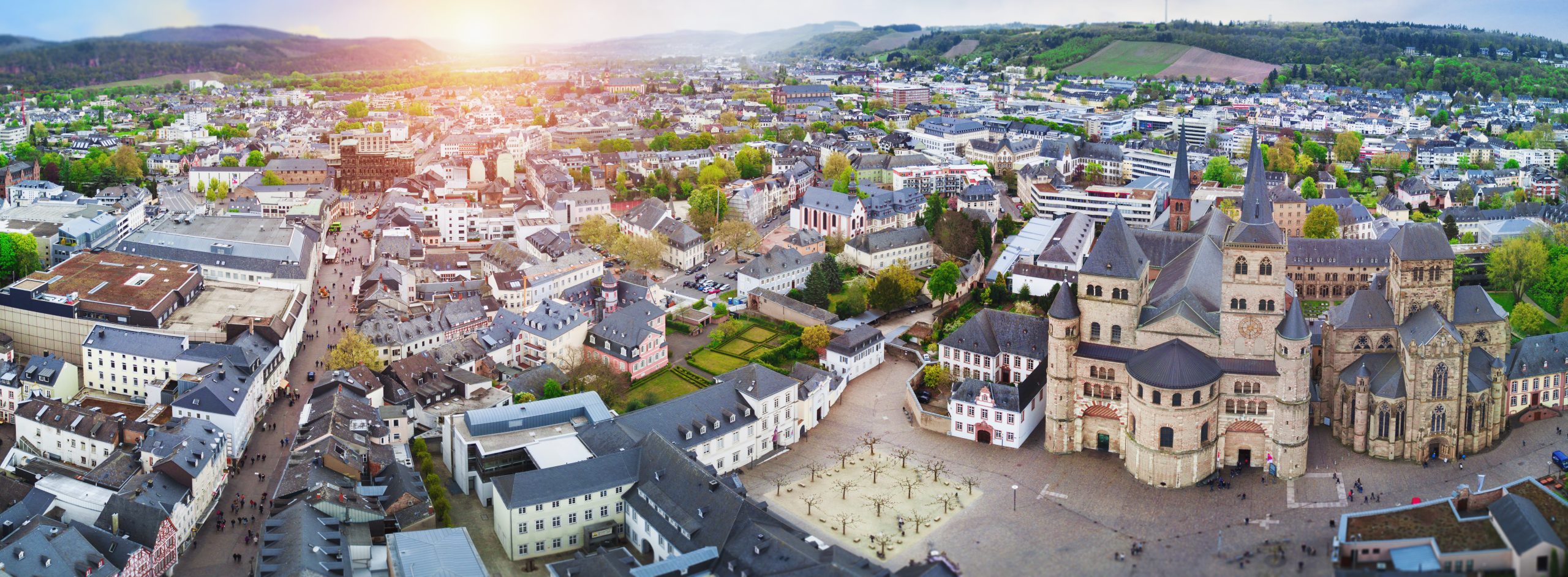 Panoramaaufnahme der Innenstadt von Trier mit dem Dom im Vordergrund und der historischen Altstadt im Sonnenlicht. Im Hintergrund sind grüne Hügel und die Moselregion zu erkennen.
