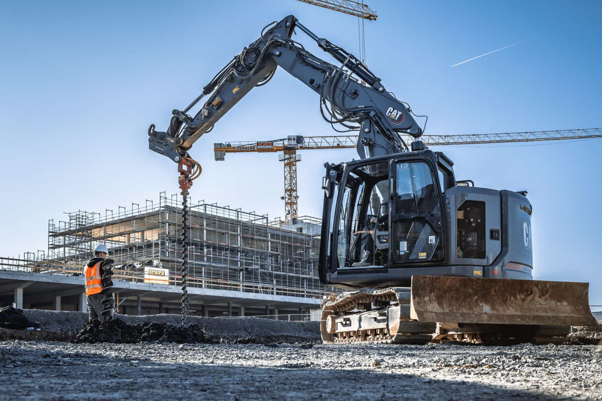 Bagger führt auf einer Baustelle Bohrarbeiten durch, im Hintergrund stehen Kräne und ein im Bau befindliches Gebäude.