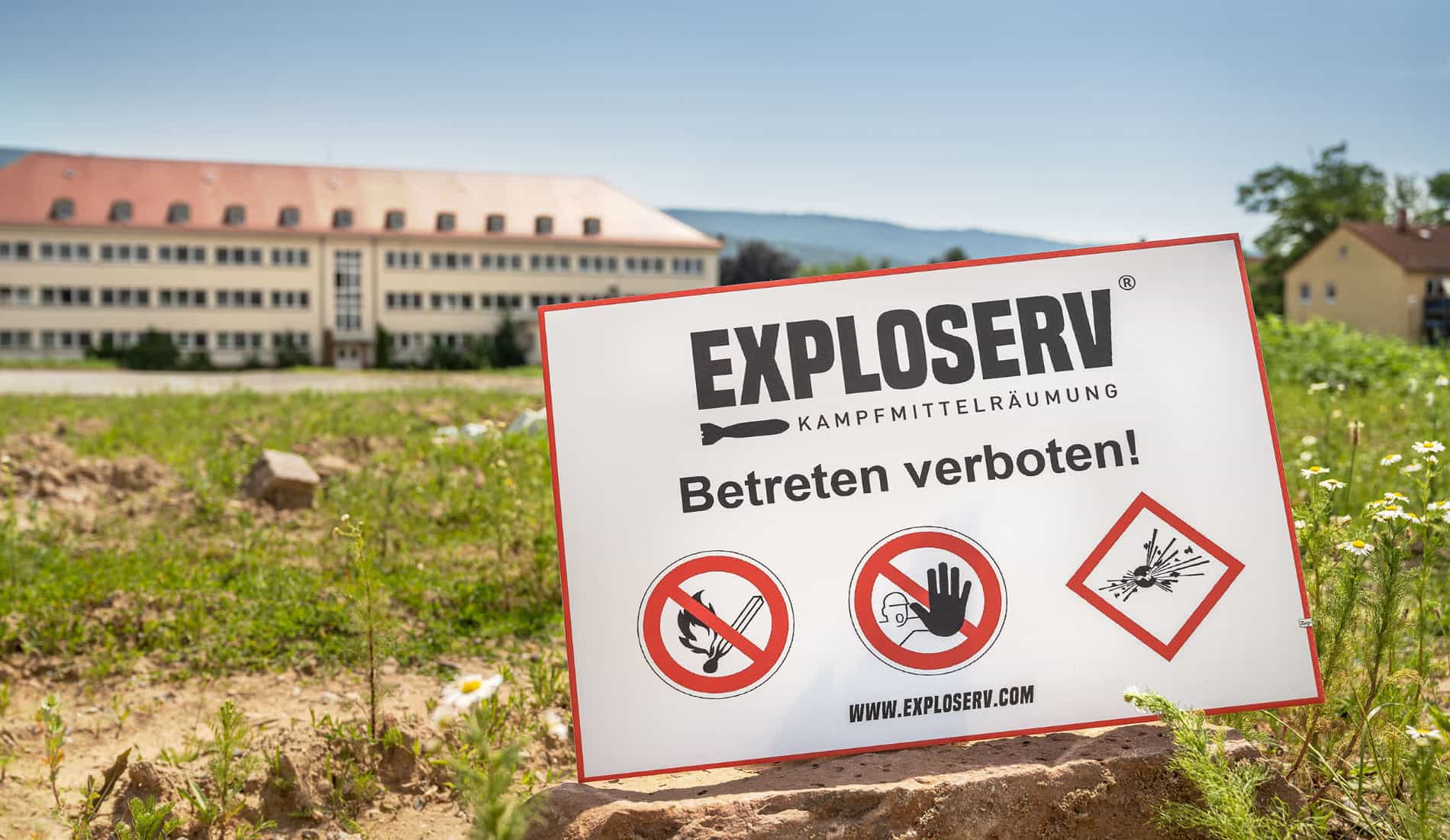 Warnschild mit der Aufschrift „EXPLOSERV – Kampfmittelräumung. Betreten verboten!“ steht auf einer abgesperrten Wiese, im Hintergrund ein Gebäude.