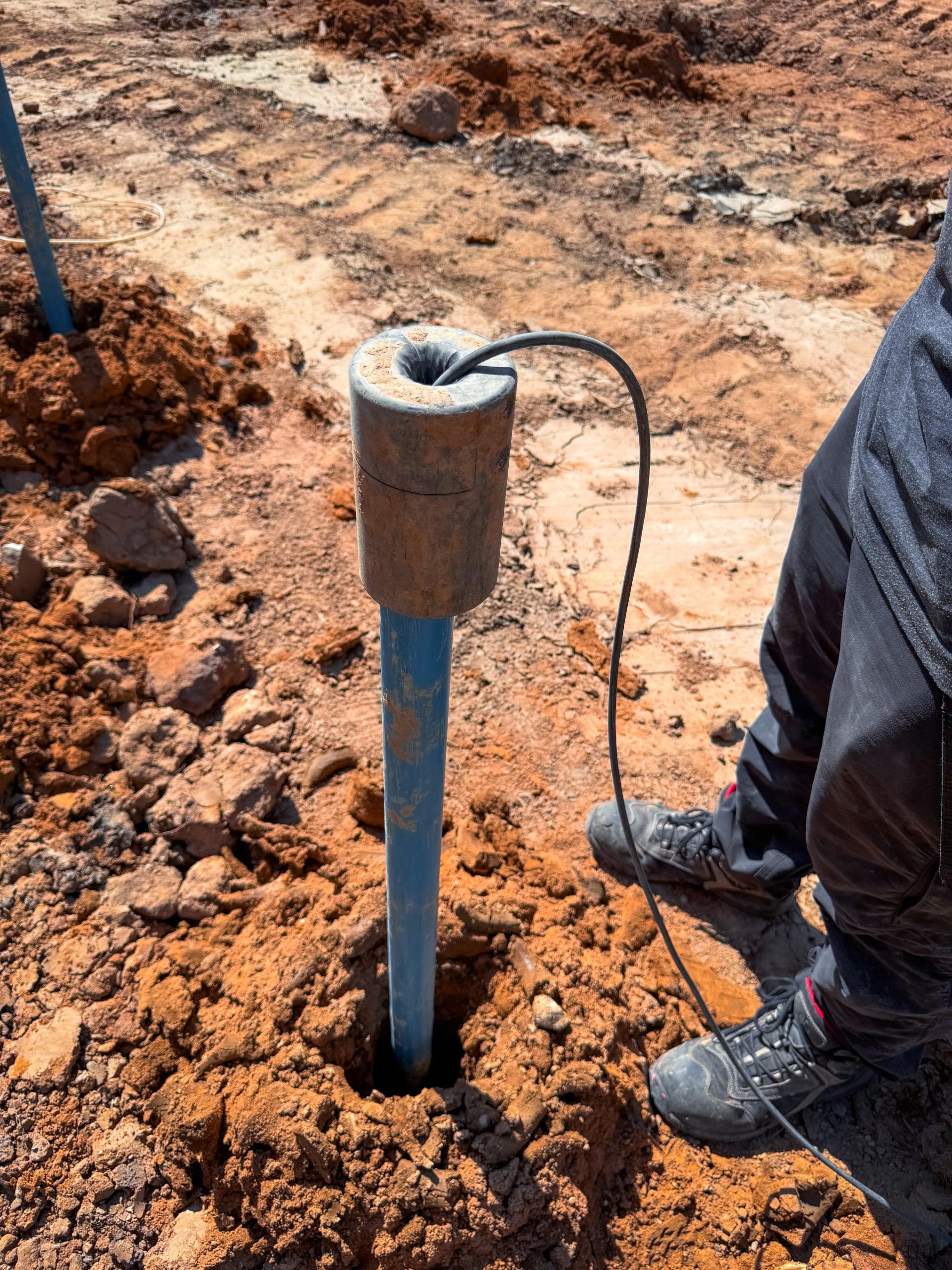 Person steht auf einer Baustelle neben einem in den Boden eingesetzten blauen Rohr mit Messsonde und Kabel. Der Boden besteht aus rötlicher Erde.