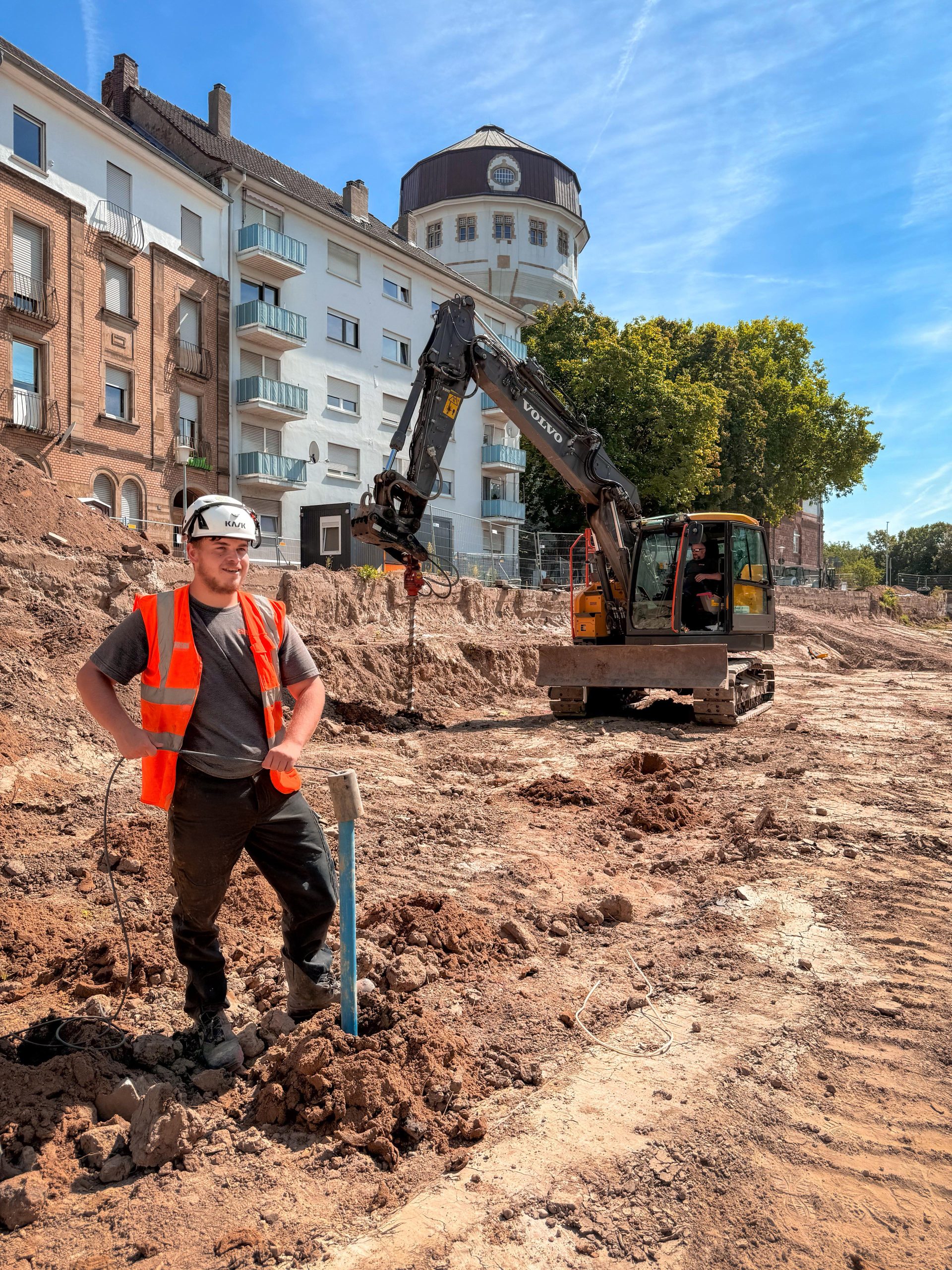 Bauarbeiter mit Schutzhelm und orangefarbener Warnweste steht auf einer Baustelle neben einem Messrohr. Im Hintergrund arbeitet ein Bagger, dahinter Wohngebäude und ein Wasserturm.