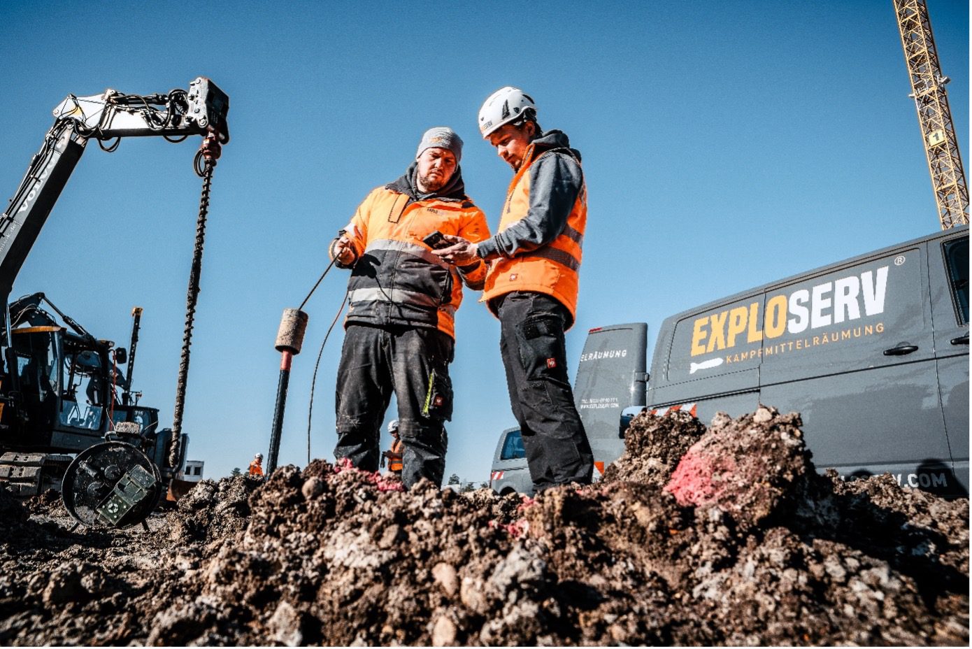Zwei Arbeiter mit Schutzhelmen und orangefarbenen Warnjacken stehen auf einer Baustelle und prüfen gemeinsam Messdaten. Neben ihnen steht ein Bagger, im Hintergrund ist ein Container mit dem Firmenlogo ‚Exploserv‘ zu sehen.
