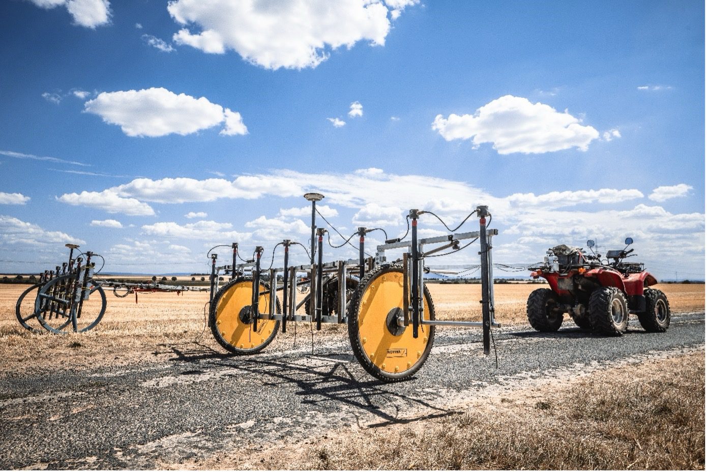 Ein rotes Geländefahrzeug zieht eine großflächige Messvorrichtung mit mehreren gelben Rädern über ein trockenes Feld. Der blaue Himmel mit weißen Wolken betont die Weite der Landschaft und den technischen Einsatz im Freien.